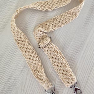 May include: A cream-colored macrame bag strap featuring a textured, knotted cord design. The strap includes a silver-toned metal adjustable slider buckle and two silver-toned swivel lobster claw clasps at each end for secure attachment.