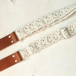 May include: Cream-colored macrame guitar strap with brown leather ends and silver-tone hardware. The strap features a woven geometric pattern and adjustable buckle. A stylish accessory for musicians.