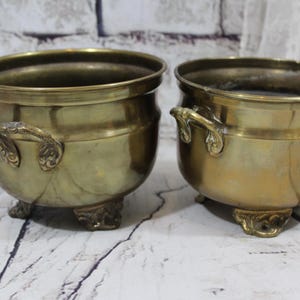 May include: Two antique brass plant pots with ornate handles and feet. The pots are both round and have a slightly distressed finish.
