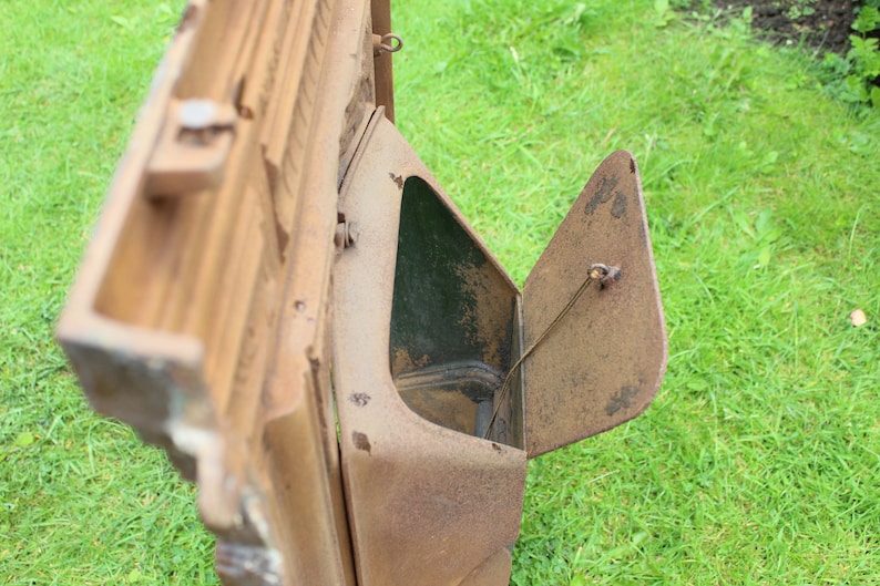 Pu&ograve; includere: Un inserto per camino in metallo arrugginito con una porta a cerniera. La porta &egrave; aperta, rivelando un interno scuro. L'inserto &egrave; seduto su un letto di erba verde.