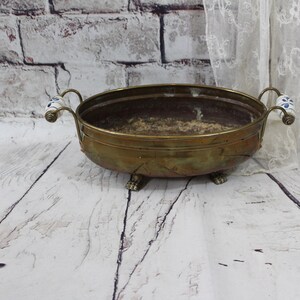 May include: An oval brass bowl with a weathered patina, featuring two curved handles with white and blue floral ceramic accents. The bowl is supported by four small, ornate metal feet and has a deep, open interior basin with a dark, textured base.