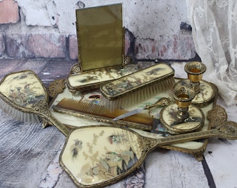 Può includere: Un set da toeletta vintage con finitura dorata. Il set comprende due specchietti da mano, due portacandele, un pettine, una spazzola e una piccola cornice. Gli specchi e la spazzola sono decorati con scene dipinte di persone e uccelli.