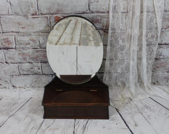 Può includere: Uno specchio da toeletta vintage in legno con uno specchio ovale e un piccolo scomparto di stoccaggio. Lo specchio ha una cornice in legno scuro e lo scomparto di stoccaggio ha un coperchio incernierato.