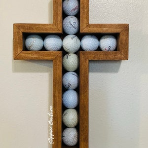 May include: A wooden cross with a golf ball holder. The cross is stained a dark brown color and has a rectangular opening in the center. The opening is filled with golf balls, which are arranged in a grid pattern. The cross is mounted on a white wall.