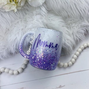 May include: White ceramic mug with a purple glitter ombre design and the word "Momma" written in purple cursive. The mug is on a white faux fur surface.