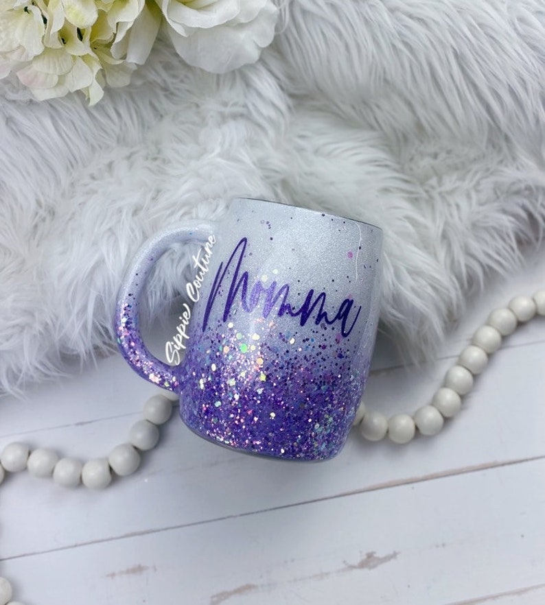 May include: White ceramic mug with a purple glitter ombre design and the word "Momma" written in purple cursive. The mug is on a white faux fur surface.