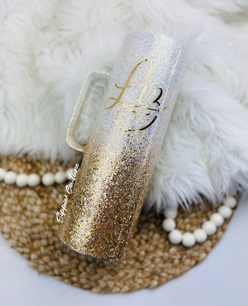 May include: A gold and white glitter tumbler with a handle. The tumbler has the name "Liz" written on it in gold lettering.
