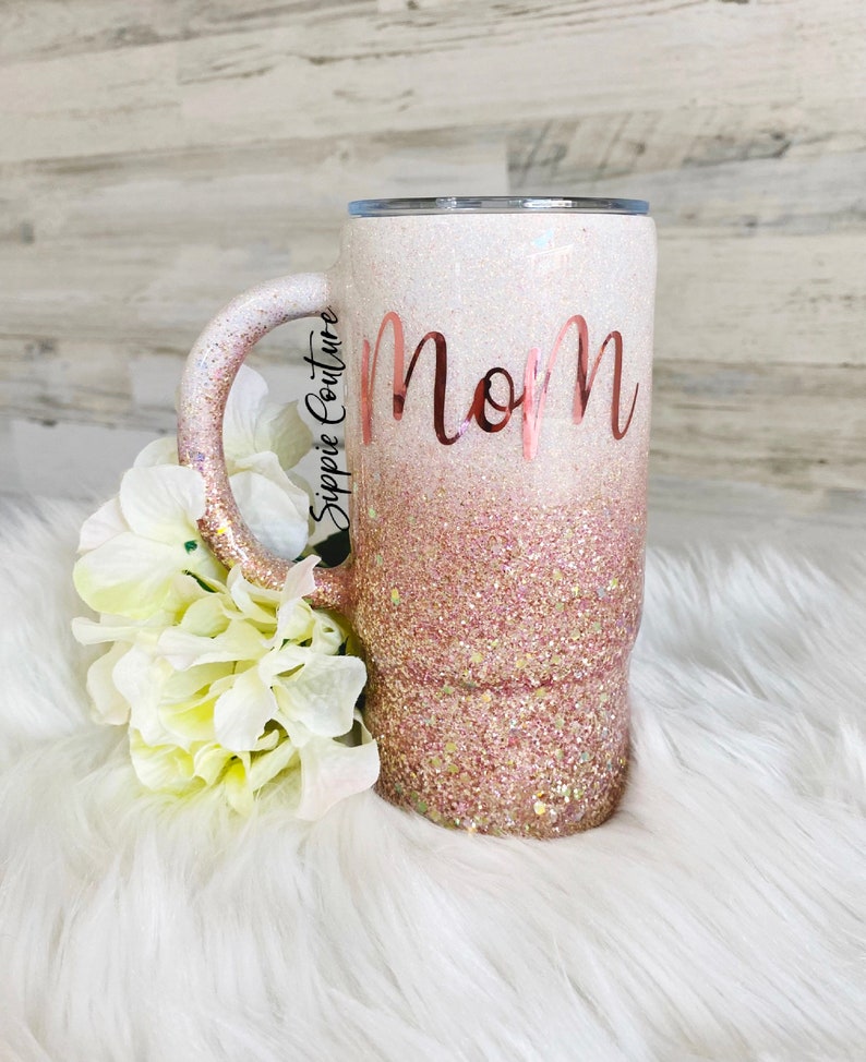 May include: A white and pink glitter tumbler with a rose gold handle and the word "Mom" in rose gold lettering.