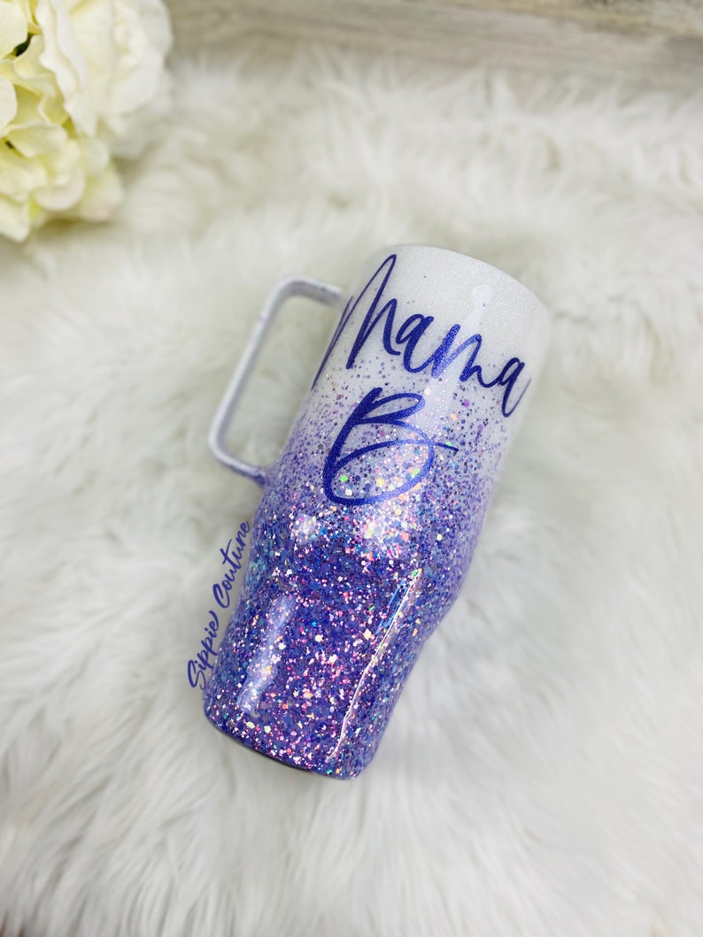 May include: A white and purple glitter tumbler with a handle. The tumbler has the text "Mama B" written on it in purple.
