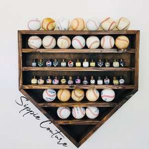 May include: A wooden baseball diamond-shaped shelf with four shelves. The top shelf has 10 baseballs, the second and third shelves have small bottles, and the bottom shelf has 6 baseballs. The shelf is mounted on a white wall and has the text "Sippie Couture" written on the bottom right corner.