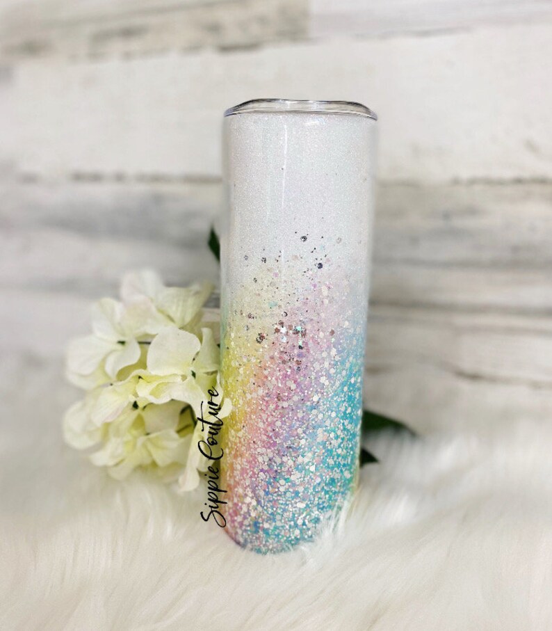 May include: A white tumbler with a rainbow glitter ombre design. The tumbler has a clear lid and is decorated with the text "Sippie Couture".