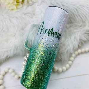 May include: A white and green glitter tumbler with a handle. The tumbler has a white top with green glitter fading to a darker green at the bottom. The word "Mermaid" is written in green cursive on the white part of the tumbler.