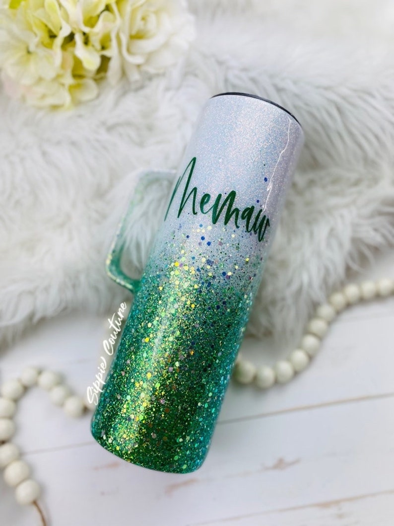 May include: A white and green glitter tumbler with a handle. The tumbler has a white top with green glitter fading to a darker green at the bottom. The word "Mermaid" is written in green cursive on the white part of the tumbler.