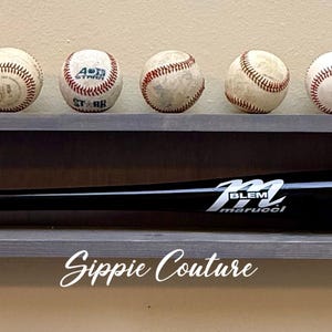 May include: A gray wooden wall-mounted display shelf holding a black baseball bat with white text reading M Marucci Lakeshore Titan 2025 Mason Maldonado #11. Nine white baseballs with red stitching are lined up along the top edge of the shelf.