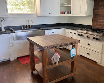 May include: A rustic wooden kitchen island with three shelves and wheels. The island has a dark brown stain and is made of reclaimed wood. It has a metal bar with two hooks on the side. A red oven mitt hangs on one of the hooks.