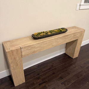 May include: A light-colored wooden bench with a rectangular shape. The bench has a natural wood grain and a dark wooden bowl filled with moss and decorative elements. The bench is placed against a light-colored wall and dark wood floor.