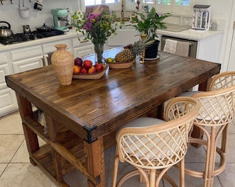 May include: A rustic wooden kitchen island with a dark brown stain. The island has a built-in shelf and is topped with a thick wooden countertop. Two woven rattan bar stools with cream-colored cushions are placed next to the island.