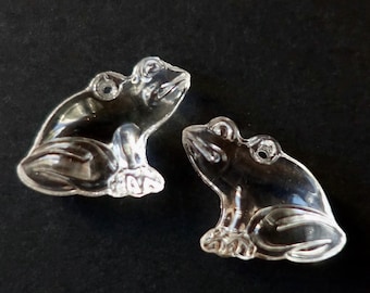 May include: Two clear, transparent frog figurines are displayed against a black background. The frogs are detailed with visible eyes, legs, and a textured surface, suggesting a glass or crystal material. The frogs are positioned facing each other.