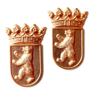 Könnte beinhalten: Zwei kupferfarbene Metallabzeichen. Jedes zeigt einen Bären, der auf seinen Hinterbeinen in einem schildförmigen Feld steht, das von einer Krone gekrönt wird. Wahrscheinlich dekorative oder symbolische Artikel.