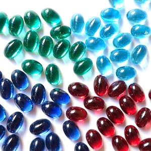 Könnte beinhalten: Eine Ansammlung farbiger, oval geformter Glasperlen in Grün-, Blau- und Rottönen. Die Perlen sind auf einer weißen Oberfläche verstreut, wobei jede Farbe gruppiert ist. Die durchscheinenden Perlen haben eine glänzende Oberfläche.