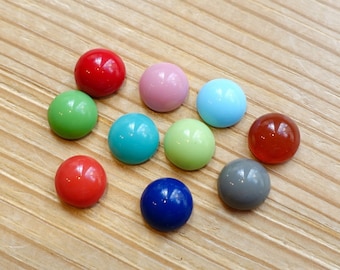 May include: Assortment of ten round, colorful cabochons. The cabochons are arranged in a semi-circle on a wooden surface. The colors include red, pink, light blue, green, turquoise, light green, orange, gray, and dark blue.
