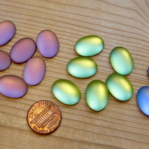 Può includere: Assortimento di cabochon ovali in vetro di vari colori, tra cui viola, blu e verde. I cabochon sono disposti in file su una superficie di legno.