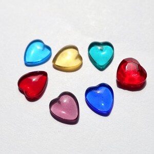 Könnte beinhalten: Sieben herzförmige Glasperlen in verschiedenen Farben, darunter Rot, Blau, Gelb und Lila. Die Perlen sind in einem Halbkreis auf einer weißen Oberfläche angeordnet.