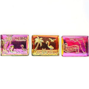 May include: Three rectangular Florida-themed magnets with a metallic finish. Each magnet features a different design: palm trees, a flamingo, and the state outline. The magnets are in shades of pink, orange, and gold.