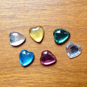 Könnte beinhalten: Sechs herzförmige Glasperlen in verschiedenen Farben, darunter klar, gelb, blau, grün und rosa. Die Perlen sind in einem Kreis auf einer Holzoberfläche angeordnet.