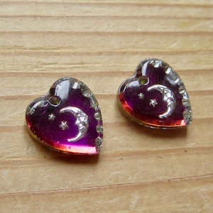 Puede incluir: Dos abalorios de vidrio en forma de corazón con un diseño de luna creciente y estrellas. Los abalorios son de color morado oscuro con un acabado de purpurina plateada.