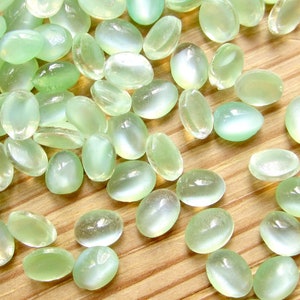 Könnte beinhalten: Eine Sammlung ovaler, hellgrüner Edelsteine mit Perlmutt-Glanz. Die Steine sind auf einer Holzoberfläche verstreut.