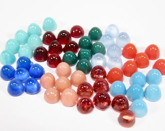 May include: Assortment of colourful cabochon gemstones in various colours, including red, blue, green, turquoise, and peach. The gemstones are arranged in a loose cluster on a white surface.