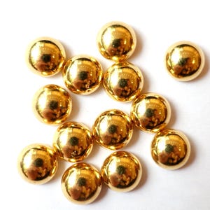 Könnte beinhalten: Eine Ansammlung glänzender, goldener, halbkugelförmiger Perlen. Die Perlen sind auf einer weißen Oberfläche angeordnet, reflektieren das Licht und erzeugen eine helle Darstellung. Diese Perlen werden wahrscheinlich für Bastelarbeiten oder Schmuck verwendet.
