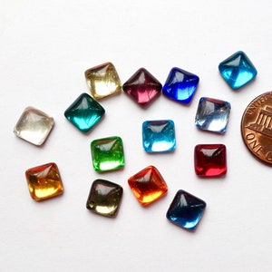 Könnte beinhalten: Eine Sammlung kleiner, quadratischer Glasperlen in verschiedenen Farben, darunter klar, gelb, grün, rot und blau. Ein Cent-Stück dient zum Größenvergleich. Die Perlen sind wahrscheinlich für Bastelarbeiten oder die Schmuckherstellung gedacht.