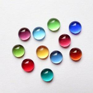 Può includere: Dieci gemme rotonde di vetro in varie tonalità di rosso, blu, verde e giallo. Le gemme sono disposte a forma di cuore su uno sfondo bianco.
