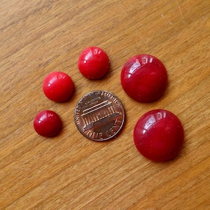 Peut inclure: Plusieurs boutons ronds rouges brillants de différentes tailles sont disposés sur une surface en bois. Une pièce de monnaie est incluse pour l'échelle. Les boutons ont une finition lisse et réfléchissante, et la couleur est un rouge vif et uni. Les boutons mesurent environ 1,2 cm à 2,5 cm de diamètre.