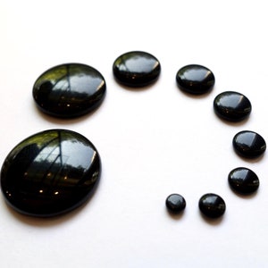 May include: A collection of black, round, smooth, polished stones of varying sizes arranged in a semi-circle on a white surface.
