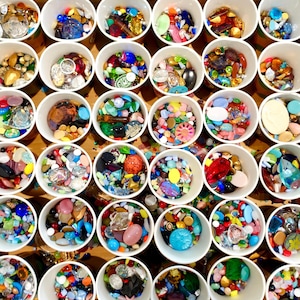 Könnte beinhalten: Sortiment an bunten Glasperlen in kleinen weißen Bechern. Die Perlen sind in verschiedenen Formen und Größen, darunter rund, quadratisch und sternförmig.