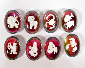 May include: Eight oval cabochons featuring white raised zodiac symbols set against a translucent deep red background with an iridescent metallic border. The symbols include a lion, ram, scorpion, fish, archer, goat-fish, maiden, and crab.