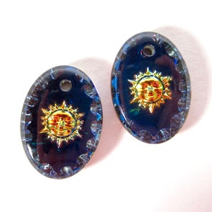 May include: Two oval-shaped glass charms featuring a gold sun face design. The charms have a dark blue iridescent finish and a hole at the top for hanging.