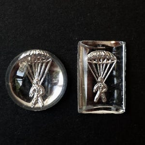 May include: Two clear glass cabochons, one circular and one rectangular, each featuring a silver-colored metallic inlay of a person descending with an open parachute. The designs are embedded within the transparent material, creating a three-dimensional effect.