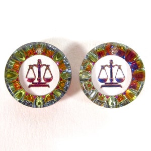 May include: A pair of iridescent earrings with a scale of justice design. The earrings are round and have a multi-colored, rainbow-like finish.