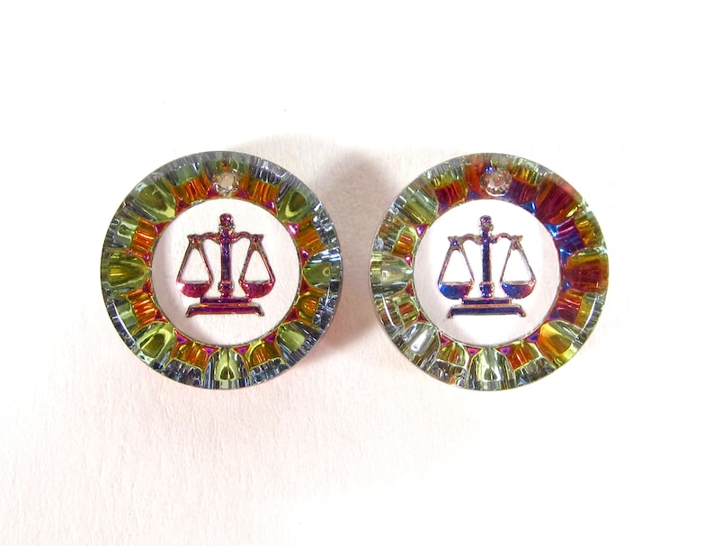May include: A pair of iridescent earrings with a scale of justice design. The earrings are round and have a multi-colored, rainbow-like finish.