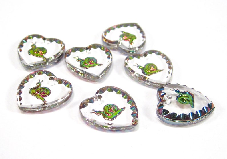 May include: Seven iridescent heart-shaped glass charms with a snail design. Each charm has a scalloped edge and a clear background.