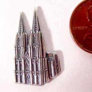May include: Silver-toned metal charm of Cologne Cathedral, a detailed miniature replica of the famous German landmark. The charm is about the size of a US penny.
