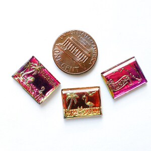 May include: Three rectangular, iridescent glass tiles with Florida-themed designs, including palm trees and flamingos, next to a US penny. The tiles have a mix of red, gold, and purple hues.