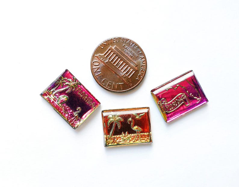 May include: Three rectangular, iridescent glass tiles with Florida-themed designs, including palm trees and flamingos, next to a US penny. The tiles have a mix of red, gold, and purple hues.