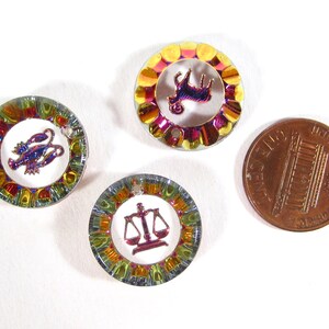 May include: Three small, round, iridescent metal charms with zodiac symbols. The charms are about the size of a US penny. The symbols are a ram, a crab, and a scale.