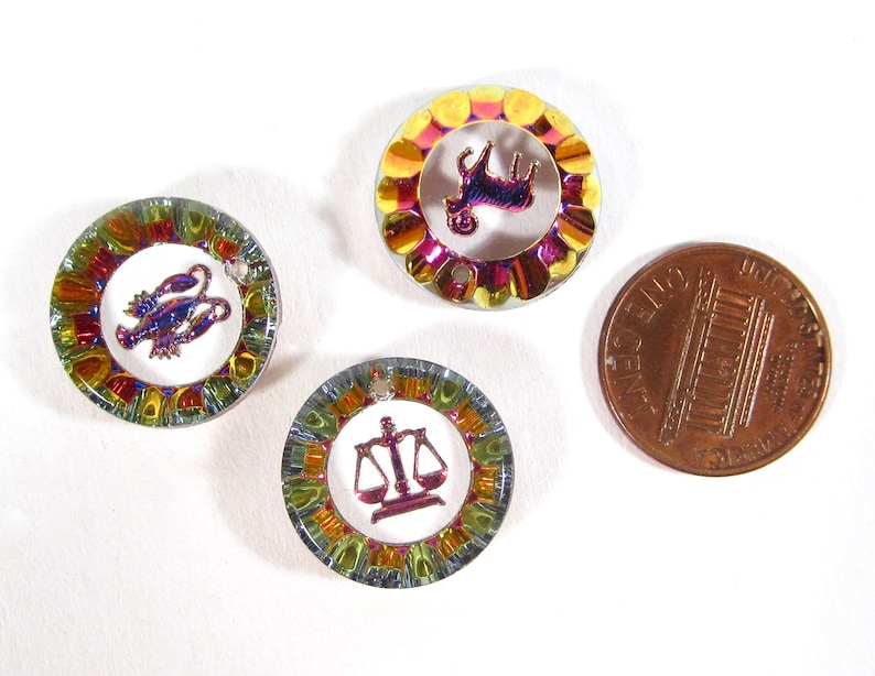 May include: Three small, round, iridescent metal charms with zodiac symbols. The charms are about the size of a US penny. The symbols are a ram, a crab, and a scale.
