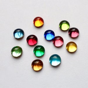 Könnte beinhalten: Eine Ansammlung farbenfroher, runder Glasperlen in verschiedenen Farbtönen wie Rot, Blau, Grün und Gelb. Die Perlen sind auf einer weißen Oberfläche verstreut, wobei sich das Licht in ihnen spiegelt. Ideal für Bastelarbeiten.